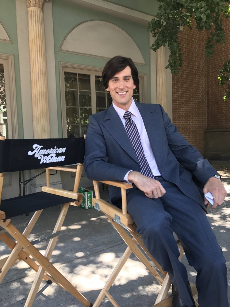 Jonathan in a suit, seated in an American Woman director's chair