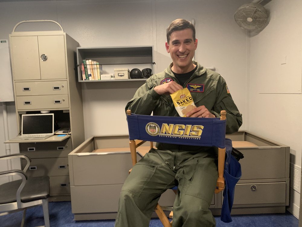 Jonathan in a flight suit, seated in an NCIS director's chair